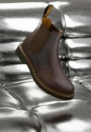Botte Chelsea en cuir marron avec coutures jaunes et panneaux latéraux élastiques reposant sur une surface capitonnée argentée brillante.