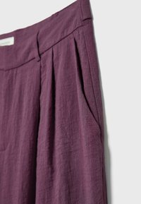 Close-up of purple linen pants showing waistband, pleats, and side pocket on a plain light background.