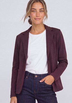 Donna con capelli chiari raccolti in uno chignon morbido, indossa una camicia bianca, un blazer viola scuro e jeans blu scuro con una mano in tasca.