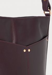 Bolso de cuero burdeos con textura suave, diseño geométrico, detalles en dorado y un bolsillo exterior.