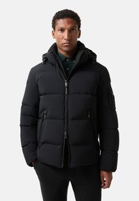 Zwarte pufferjack met een gestikte ontwerpe, rits voorkant en afneembare capuchon. Bevat zij- en mouwenzakken, gestructureerde stof en een slank silhouet.