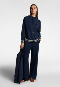 Tuta navy con felpa con zip e cappuccio regolabile, due tasche frontali e polsini a righe, abbinata a pantaloni a gamba larga e scarpe marroni.