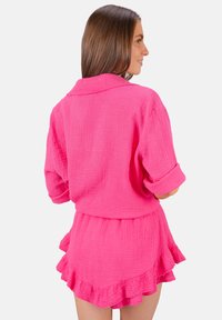 Outfit rose texturé comprenant un haut à boutons avec des manches retroussées et une jupe à volants. Le tissu semble léger et respirant.