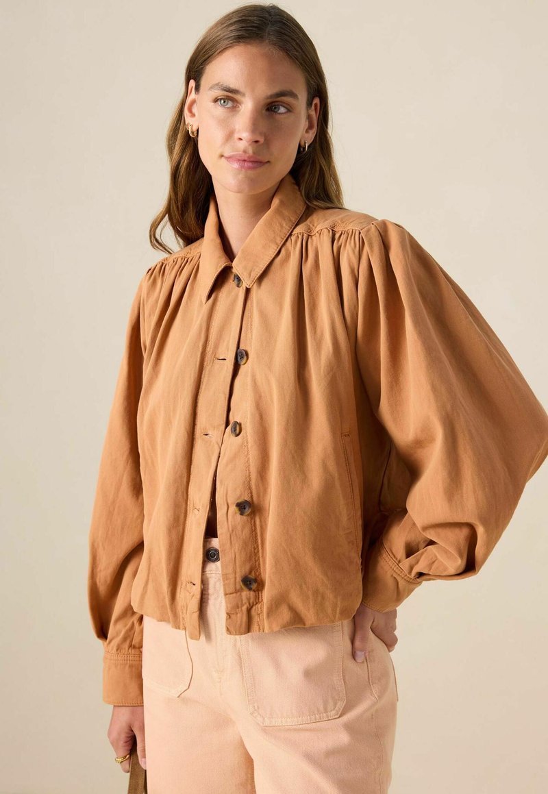 Veste en coton marron clair à manches bouffantes avec un col et une fermeture à boutons, associée à un pantalon pêche clair avec des poches avant.