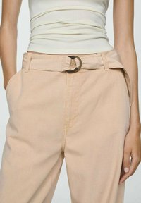 Pantaloni beige a vita alta con design con cintura, dotati di una fibbia rotonda marrone, tasche laterali e una morbida texture di tessuto liscia.