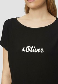 Vrouw in een zwart t-shirt met ronde hals waarop in het midden voorop witte "s.Oliver" tekst is gedrukt.
