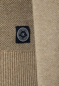 Beżowa dzianinowa tkanina swetra z teksturowanymi sekcjami, z czarną metką z białym okrągłym logo i tekstem, przyszytą na dolnej krawędzi.