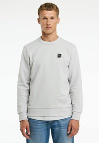 Sudadera gris claro hecha de tejido texturizado, con cuello redondo y un pequeño parche de logo negro en el frente. Combinada con jeans azules.