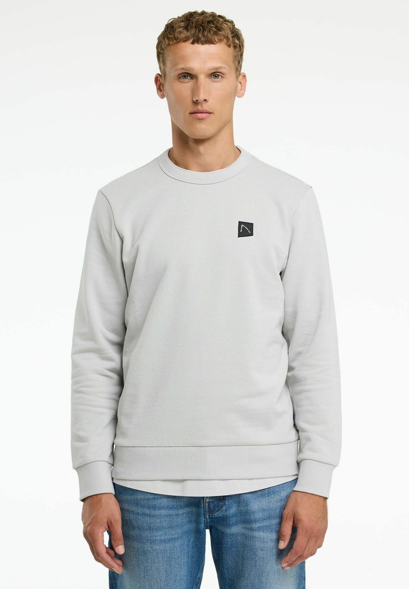 Sudadera gris claro hecha de tejido texturizado, con cuello redondo y un pequeño parche de logo negro en el frente. Combinada con jeans azules.