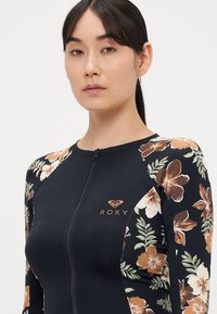 Marineblauw rashguard met lange mouwen, bloemmotief in oranje en groen op de schouders, ritssluiting aan de voorkant en een gouden Roxy-logo op de borst.