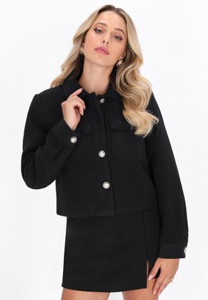 Schwarze kurz geschnittene Jacke aus strukturiertem Stoff mit zwei frontalen Taschen, silbernen Knopfverschlüssen und einem spitzen Kragen.