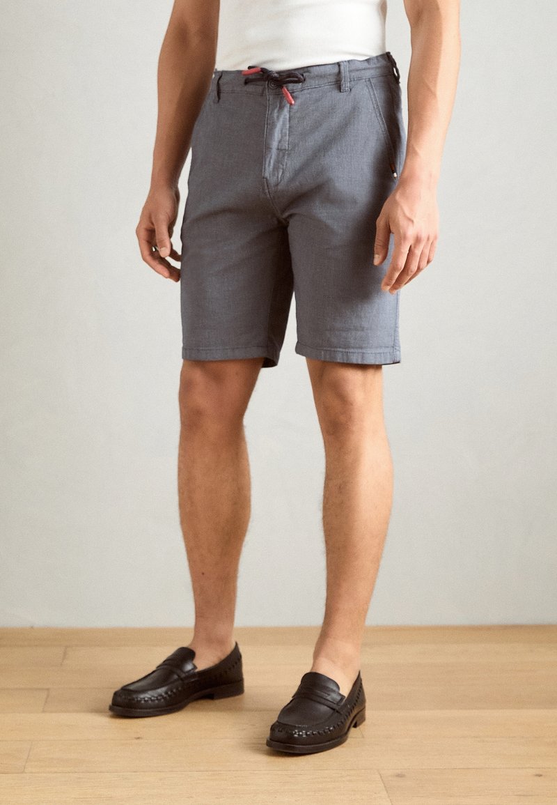 Shorts gris en tissu léger, dotés d'une ceinture noire avec un cordon de serrage rouge et de poches latérales, associés à des mocassins noirs.