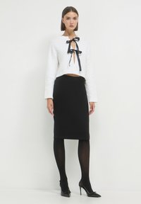 Frau in einem weißen, gestrickten, cropped Pullover mit schwarzen Satinbändern, schwarzer Bleistiftrock, schwarzen Strumpfhosen und schwarzen High Heels, stehend.