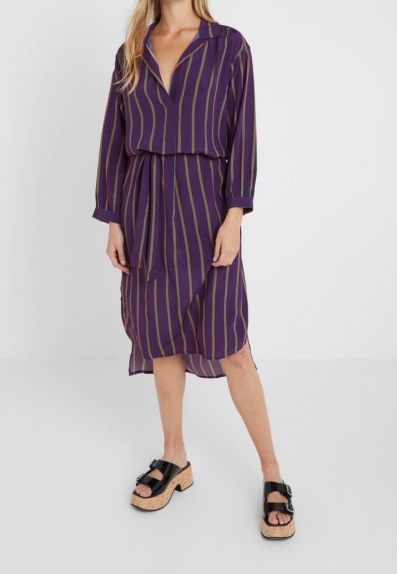 Femme portant une robe midi à rayures verticales violettes, avec col et ceinture, associée à des sandales compensées noires à semelles en liège.