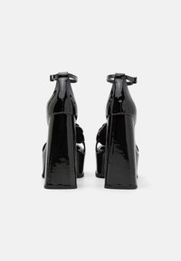 Sandales à talons hauts en cuir verni noir avec un talon bloc épais, une sangle à la cheville et un design ouvert au bout. Texture lisse et brillante avec des détails découpés.