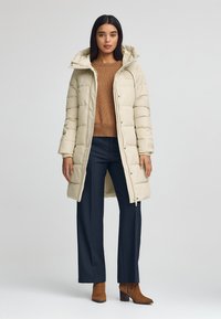 Abrigo acolchado beige con capucha, con cierre de cremallera y diseño acolchado, usado sobre un suéter marrón y pantalones azul marino. Botas de tobillo marrones completan el atuendo.