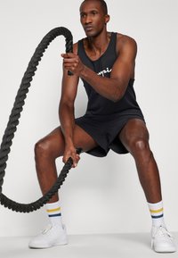Canottiera nera con logo bianco, pantaloni corti neri e sneakers bianche. Tiene una corda da battaglia nera spessa, indossando calze sportive a righe.
