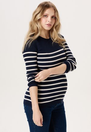 Jeune femme enceinte aux longs cheveux blonds portant un pull rayé bleu marine et blanc et un jean foncé, tenant doucement son bras.