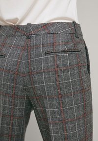 Gros plan sur un pantalon taillé à carreaux gris avec des lignes rouges et blanches, doté de passants de ceinture et de poches passepoilées à l'arrière, porté avec un haut blanc.