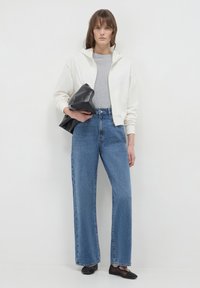 Haut de sweat zippé blanc, t-shirt gris, jean bleu à taille haute et jambes larges, ballerines noires. Modèle tenant un sac noir, debout contre un fond neutre.