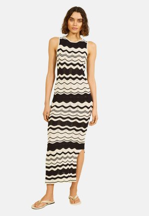 Kobieta w czarno-białej, falowanej w paski, bezrękawnikowej sukience midi z rozcięciem po boku, w połączeniu z beżowymi, płaskimi sandałami, stojąca na białym tle.