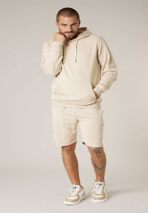 Homme debout portant un sweat à capuche beige, un short assorti et des baskets beige-blanc, avec des tatouages visibles sur les deux jambes.