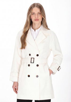 Kobieta o długich, jasnobrązowych włosach, ubrana w biały płaszcz trencz z paskiem i ciemnymi guzikami na białej koszuli, stojąca na białym tle.