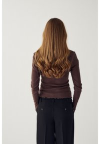 Sieviete ar gariem viļņotiem brūniem matiem, tērpusies brūnā ribotā topā un melnās biksēs, stāvot pretvienkāršu gaišu fonu.