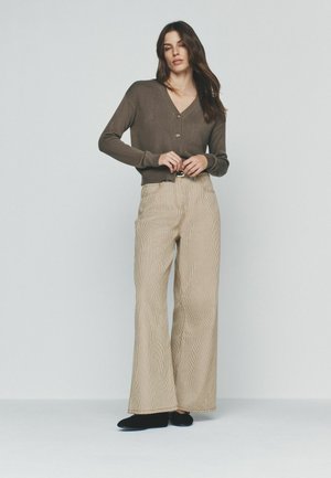 Ženska z dolgimi rjavimi lasmi, oblečena v rjav kardigan z gumbi, bež široke hlače z pasom in črne čevlje, stoji pred enobarvno ozadje.