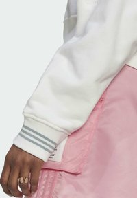 Sudadera blanca con puños rayados en gris, superpuesta a una tela rosa con una textura suave. Costuras visibles y un pequeño detalle de etiqueta.