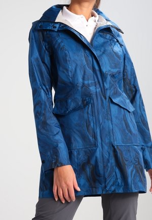 Žena nosící modrou vzorovanou nepromokavou bundu střední délky s kapsami se klopami, zipem a manžetami na druky přes bílou polokošili.