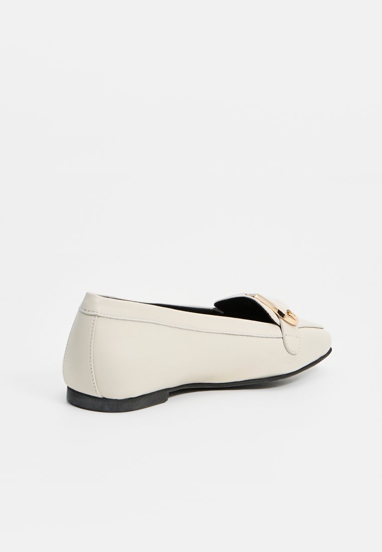 Beige Lederloafers mit abgerundetem Zehenbereich, flacher Sohle und einem goldenen Metallakzent an der Vorderseite. Glatte Textur und minimale Nähdetails.