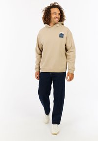 Beige hoodie met een textuurontwerp en een blauwe bergafbeelding op de linkerborst, gecombineerd met donkere denim jeans en witte sneakers.