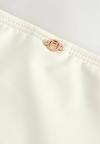 Gros plan d'un tissu crème avec une couture double piqûre et une petite boucle décorative ronde dorée fixée près du bord.