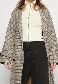 Houndstooth-patterned oversized beige and brown coat with large buttons, worn over a cream ribbed top and black jeans.