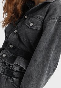 Czarna kurtka jeansowa z zapięciem na guziki z przodu, dwoma kieszeniami na piersi i luźnym krojem. Tkanina ma wyblakłą fakturę i czarny pas w talii.
