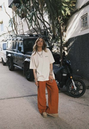 Obszerna kremowa koszulka z zielonym nadrukiem, teksturowane, szerokie spodnie w kolorze rdzy, beżowe buty wsuwane oraz wielowarstwowa srebrna biżuteria.