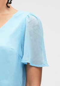 Ανοιχτό μπλε μπλούζα με V λαιμόκοψη, κοντά διάφανα φουσκωτά μανίκια και λεία υφή. Απλός σχεδιασμός με ελάχιστες ραφές.