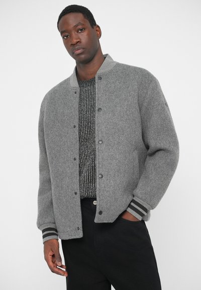 Γκρι μάλλινο μπουφάν με κουμπωμένη μπροστινή όψη, ραμμένο τελείωμα και ρίγες στους καρπούς, φορεμένο πάνω από ένα υφασμάτινο μαύρο πουλόβερ. Σκούρα παντελόνια ολοκληρώνουν το σύνολο.