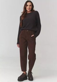 Ciemny dzianinowy sweter, brązowe spodnie z wysokim stanem i prostymi nogawkami oraz czarne, masywne buty na sznurowadła. Modelka stoi z rękami w kieszeniach.