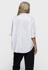 White long-sleeve shirt with a relaxed fit, featuring a back yoke, folded sleeves, and a curved hem. Soft cotton material.