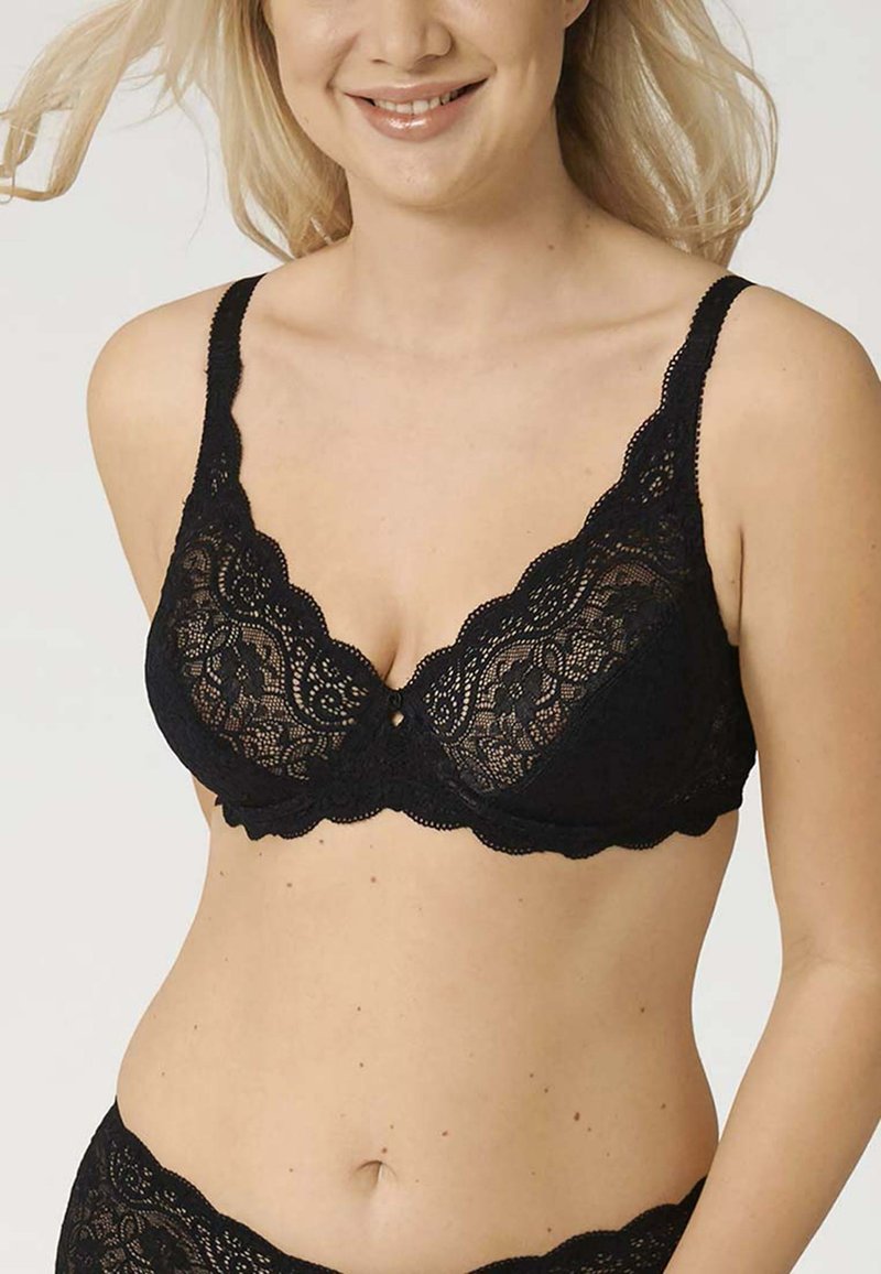 Reggiseno in pizzo nero con bordi scallopati, motivi floreali e un piccolo bottone decorativo al centro. Presenta spalline regolabili e una texture trasparente.