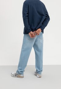 Marinblå långärmad skjorta med avslappnad passform, ljusblå jeans i denim och silverfärgade träningsskor. Händerna hopknäppta bakom ryggen, stående mot en enfärgad bakgrund.