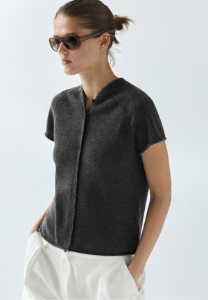 Frau mit braunen Sonnenbrillen, dunkelgrauem, kurzärmligem, geknöpftem Cardigan und weißer Hose mit Händen in den Taschen.