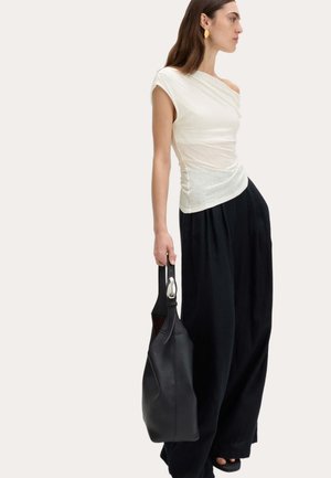 Frau in weißem Off-Shoulder-Top und weiten schwarzen Hosen, die eine schwarze Lederhandtasche mit Metallakzent vor einem einfarbigen Hintergrund hält.