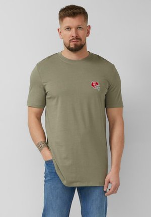 Mężczyzna w oliwkowo-zielonej koszulce z naszywką czerwonego hełmu futbolowego, niebieskich dżinsach i z tatuażem na nadgarstku, stojący na tle jednolitego szarego tła.