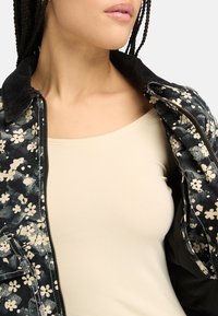Giacca nera con motivo floreale e colletto in corduroy, aperta su una maglia beige, che mette in mostra un tessuto morbido e dettagliati motivi floreali.