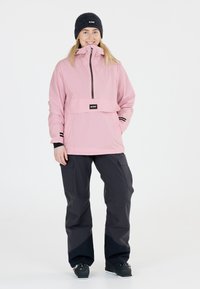 Rosa technische Jacke mit halbem Reißverschluss, Fronttasche und schwarzen Akzenten. Kombiniert mit dunklen Cargo-Hosen und einer gestrickten schwarzen Mütze.