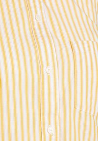 Chemise rayée jaune et blanche avec un devant à boutons, une poche poitrine et une texture de tissu lisse. Les boutons sont blancs avec un design à quatre trous.
