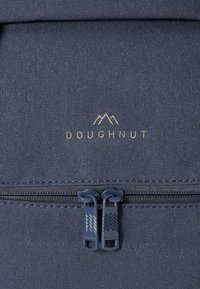Tamsiai mėlynas audinio kuprinės su užtrauktuku skyriumi, subtiliais siūlais ir auksiniu „DOUGHNUT“ logotipu su kalnų grafika priekyje.